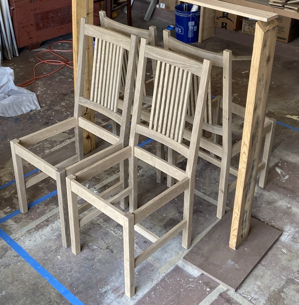 Four matching slat-back dining chairs in pale oak, unfinished, standing in the workshop